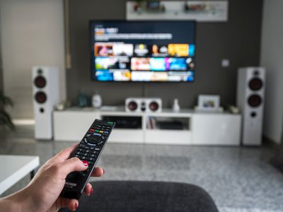 A woman's hand holding a remote control and pointing it at a TV surrounded by speakers.