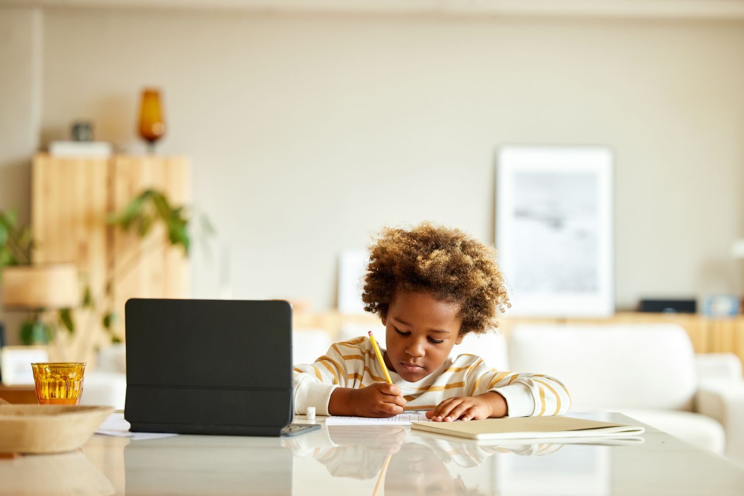 A child writing on paper with a tablet computer on the counter nearby.