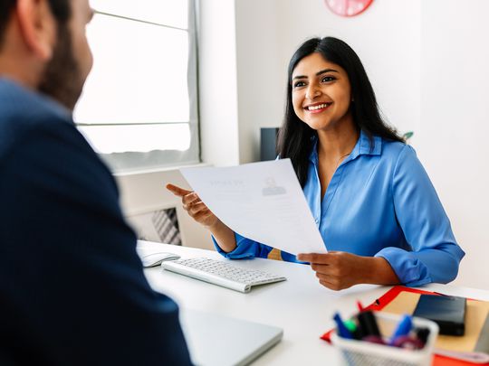 A hiring professional holding a resume and speaking with a candidate in an office setting. 