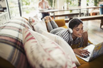 Woman lying on her stomach on a couch listening to music via headphones connected to her computer