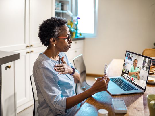 A woman sitting at a kitchen table looking at her laptop with a video call playing on it