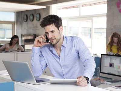 A businessman sitting at his desk and talking on his iPhone while looking at his Macbook.