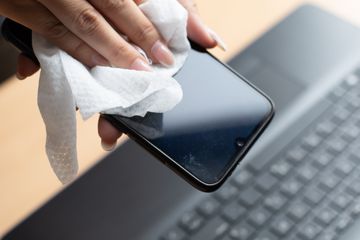 Hands using a wipe to clean a smartphone with a laptop in the background.