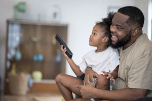 A man and his child smiling at a TV off-screen while the child holds a TV remote 